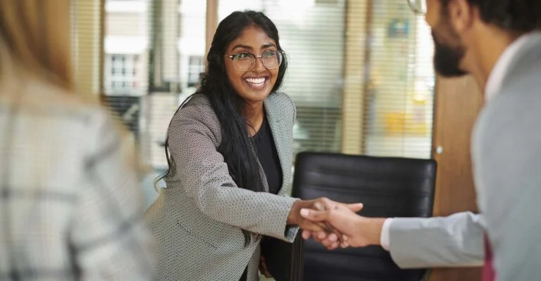 A smiling woman with long dark hair and glasses shakes hands with a man, viewed over his shoulder.