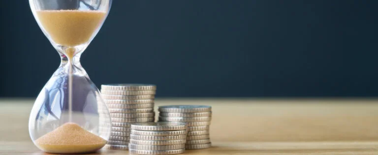 An hourglass next to three stacks of coins on a table.